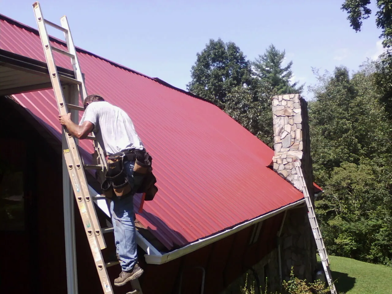 Roofer on a ladder beside a red metal roof and stone chimney — Soffit & Fascia Repair by Roofing Brothers in Castleton On Hudson
